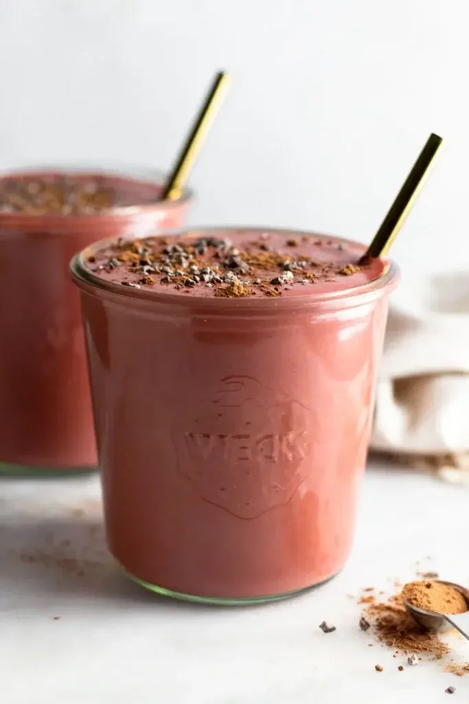 Batido de remolacha en una jarra con nibs de cacao y una pajita dentro. Delante hay una cucharilla llena de cacao en polvo. Detrás hay otro tarro lleno de batido.