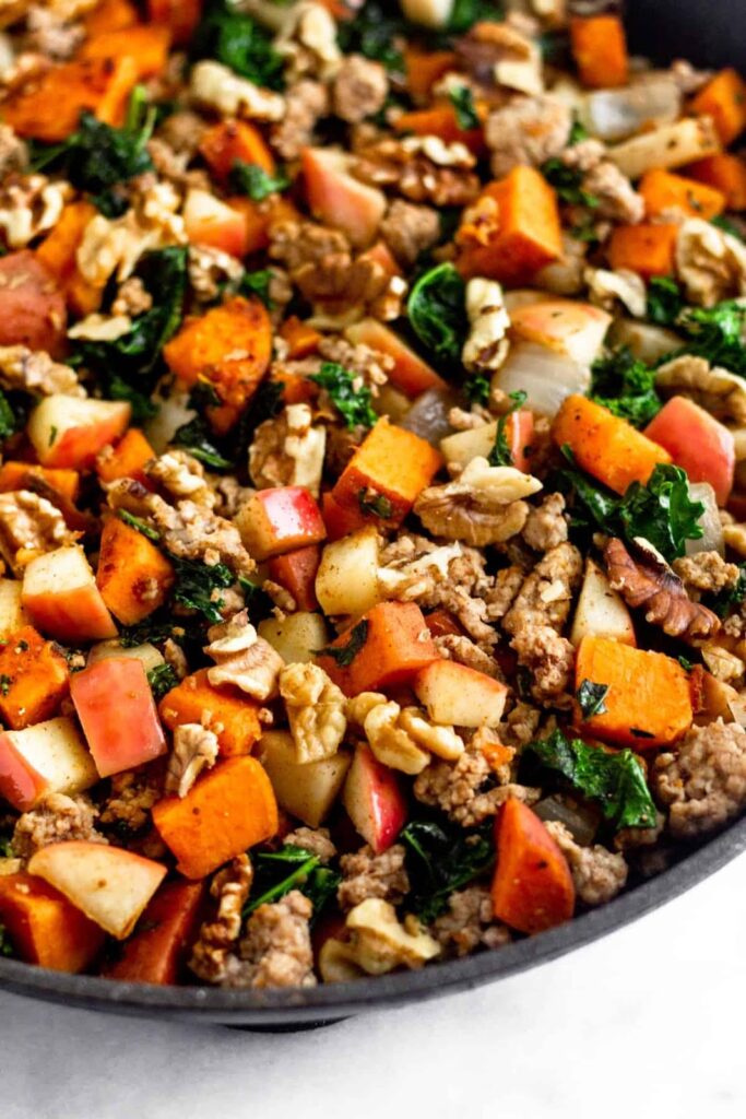 Primer plano de una gran sartén con carne picada de cerdo, boniatos, col rizada, manzanas y nueces.