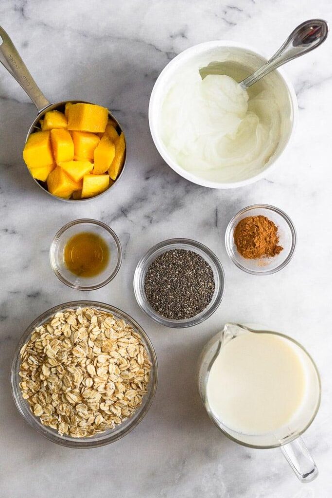 Encimera blanca con un envase de yogur griego, un vaso de leche de almendras, un bol de copos de avena, una taza de mangos cortados en dados, un bol de semillas de chía, un bol de canela y un bol de extracto de vainilla.