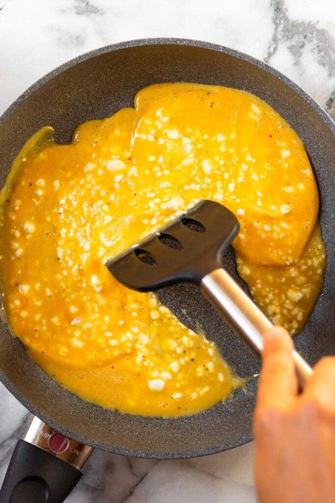 Sartén grande con huevos revueltos sin cocer. Una espátula los empuja en la sartén.