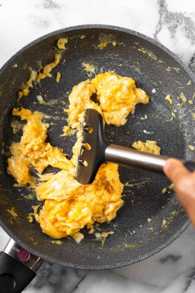 Sartén grande llena de huevos revueltos con una espátula moviéndolos mientras se cocinan.
