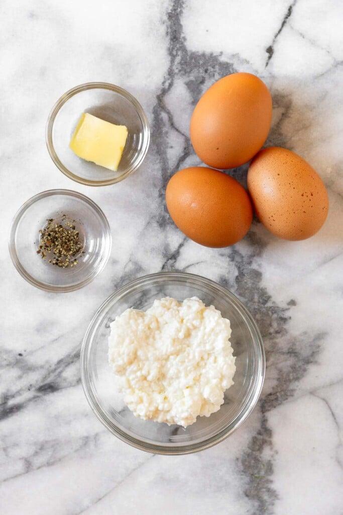Encimera de mármol blanco con tres huevos morenos, un cuenco de requesón, un pequeño cuenco de pimienta y un cuenco con una rodaja de mantequilla.