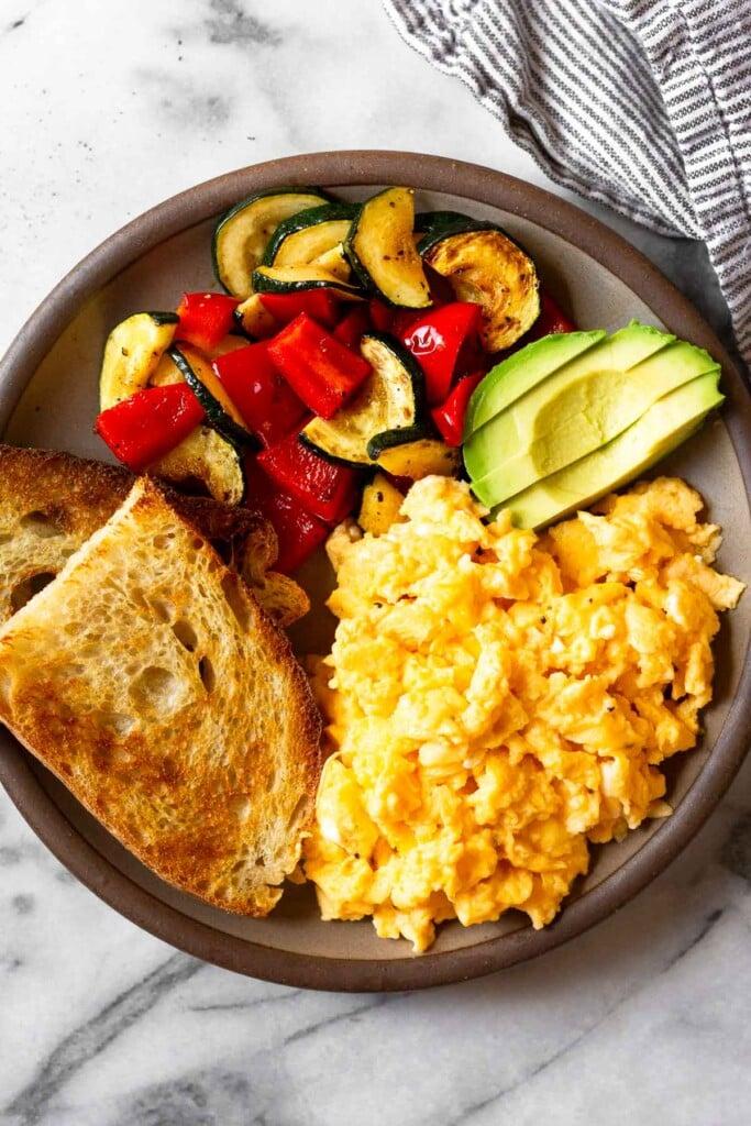Un plano general de un plato con huevos revueltos con requesón, dos tostadas, verduras salteadas y rodajas de aguacate.