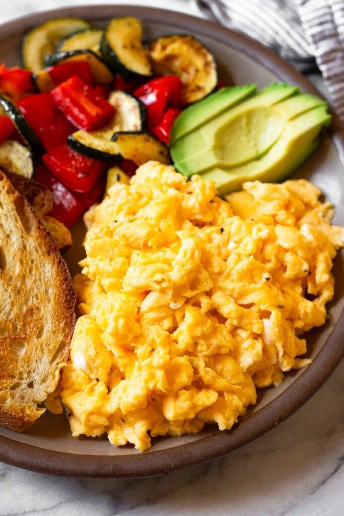 Huevos revueltos con requesón en un plato. En el plato también hay tostadas, verduras salteadas y aguacate.