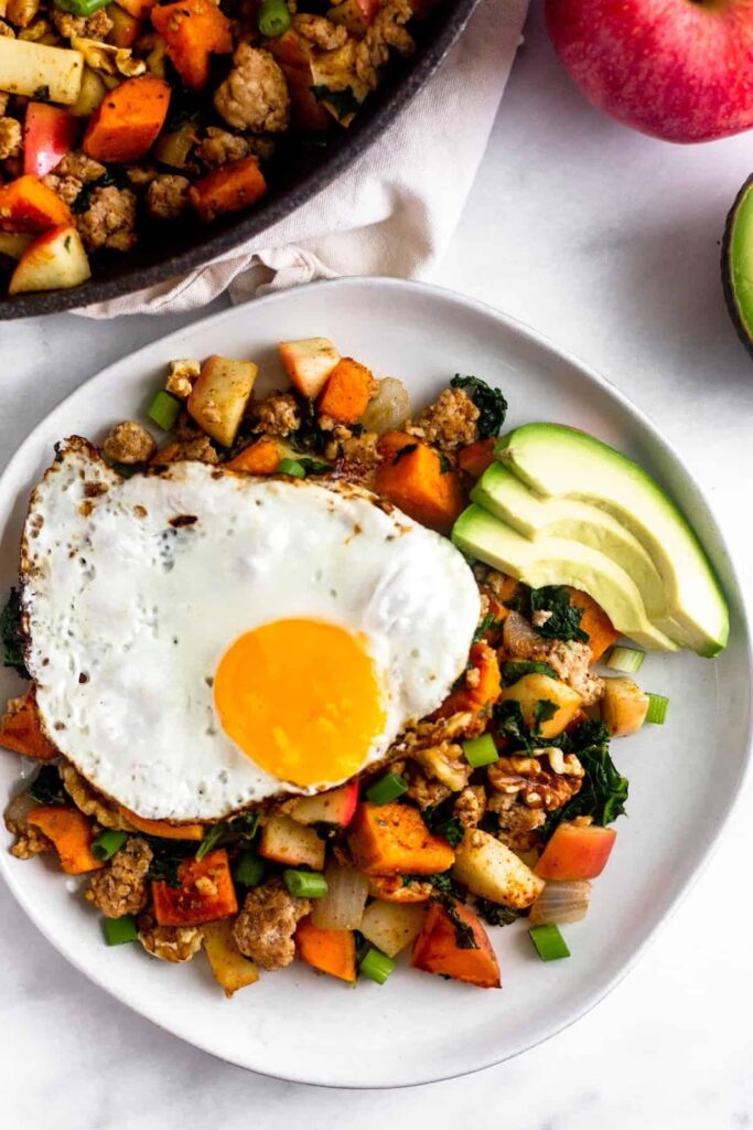 Toma aérea de un plato blanco lleno de hash paleo de boniato con salchicha y manzanas coronado con un huevo frito y aguacate. Al lado hay una sartén grande con el hash, una manzana y medio aguacate.